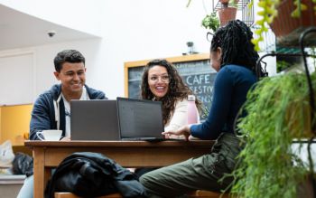 Three students chatting around a laptop in the café at Attenborough Centre for the Creative Arts (ACCA)