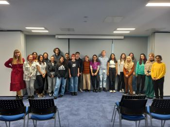 A group of Green Impact Project Assistants lined up for a photograph with smiles on their faces in front of a grey wall and chairs in front of them