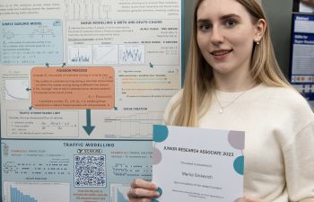 Young lady holding a certificate in front of a large poster in an exhibition hall