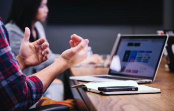 Stock image by Headway. hands gesticulating in front of a laptop screen in the foregroud with a blurred out colleague in the background