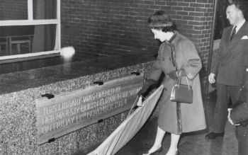 Elizabeth II removing the curtain that covered a plaque. The plaque reads this Library was opened by her Majesty Queen Elizabeth the Second on Friday 13th November 1964