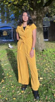 Photo of Manasi Barmecha standing on grass. Manasi is a Brown femme person, wearing a yellow jumpsuit.