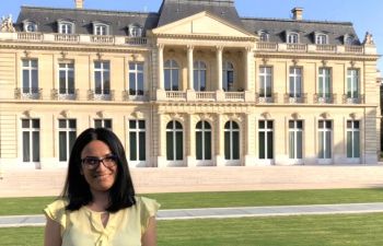 Picture of Yasemin Aslan, a woman with dark hair and a yellow top outside a building The Château de la Muette