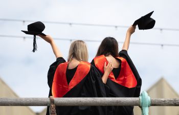 The backs of two graduates wearing graduation outfits with their arms around each other. They are posing on the Brighton beach front.