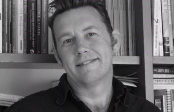 A black and white head and shoulders image of a smiling Professor Peter Newell standing in front of book shelves