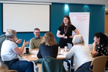 Staff sit around a table, a woman leads the conversation with her hands in the air, another stands up to write on a flipchart