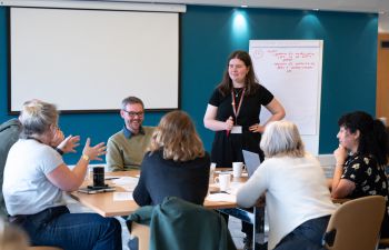 Staff sit around a table, a woman leads the conversation with her hands in the air, another stands up to write on a flipchart