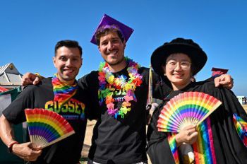 A photo of three people wearing graduation gowns with rainbow fans, and rainbow University of Sussex t-shirts. There is a blue sky in the background.