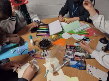 A group of individuals engaged in a craft activity around a table, with hands visible working on various projects. The table is scattered with art supplies like colored paper, markers, glue, and stickers