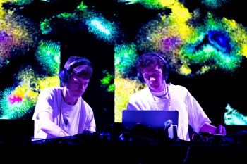 Music duo 'eyeveyes' performing on stage. Behind them is a multicolour pattern projected on a screen.