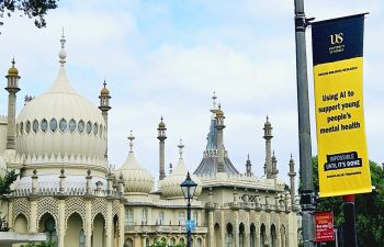 A yellow flag hangs from a lampost outside the Royal Pavilion. The text reads 'University of Sussex, Ground Breaking Research, Using AI to support young people's mental health, Impossble Unitll it's Done, Sussex.ac.uk/impossible