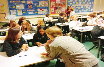 Young students seated at desks in a classroom, with a female teacher leaning down talking to two students at the front of class