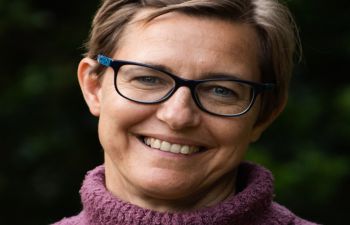 Head shot of woman with short blonde hair wearing glasses and a purple jumper and smiling to camera.