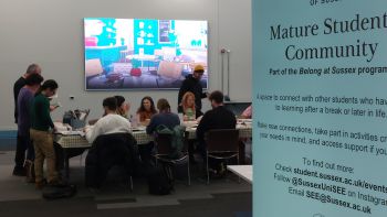 Students gather around a table. A blue sign in the foreground reads