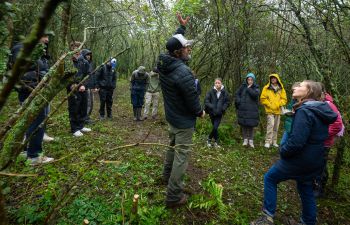 A group of students in a shrubby clearing surrounding by trees, looking at the teacher (a man in a cap) pointing and explaining