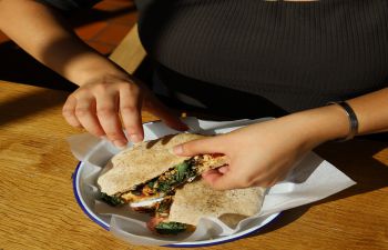 You can see the torso of a woman wearing a black top who's tearing into a healthy-looking flatbread; indoors but with the sun shining on her