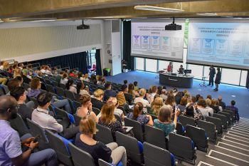 An image of audience members sitting in an auditorium with presenters at the front of the stage
