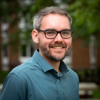 A man with greying hair and a short beard, wearing black glasses and a shirt, looking directly into the camera and smiling.