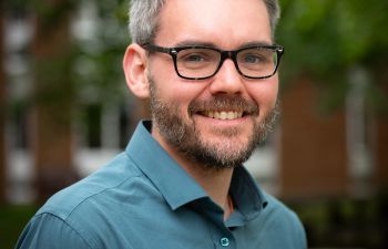 A man with greying hair and a short beard, wearing black glasses and a shirt, looking directly into the camera and smiling.