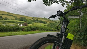 Grey bicycle in the bottom right foreground, at the side of a country road. Green fields and trees in the background.
