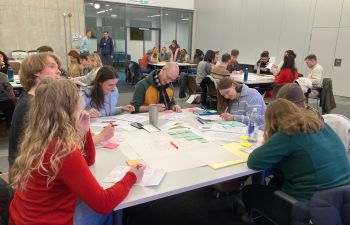 A room full of staff and students sit around tables, talking and writing with pen and paper. They are engaged in intense discussions, thinking deeply as they collaborate.