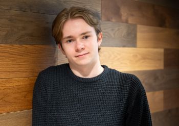 Jake Ryan smiling at the camera and leaning against a wooden wall