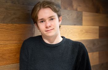 Jake Ryan smiling at the camera and leaning against a wooden wall