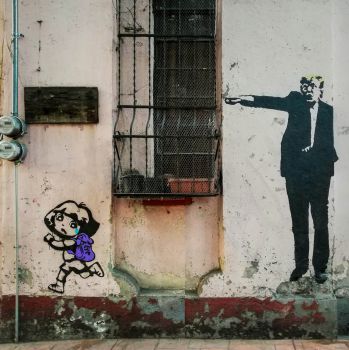Graffiti cartoon on the side of a run-down industrial building. Donald Trump is standing, wearing a suit, and pointing with his arm outstretched. Below him, Dora the Explorer (a cartoon child with backpack) is running away.