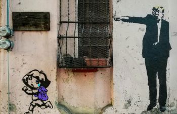 Graffiti cartoon on the side of a run-down industrial building. Donald Trump is standing, wearing a suit, and pointing with his arm outstretched. Below him, Dora the Explorer (a cartoon child with backpack) is running away.