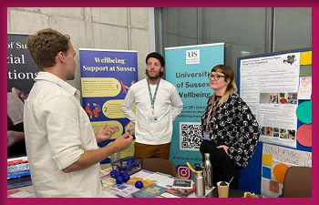 Three individuals are standing and conversing at a booth with various informational posters and materials. The booth features banners for 
