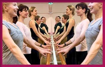 A group of people standing in front of a ballet barre, practicing ballet positions. They are facing each other in two rows, with their hands resting on the barre. The image is framed with a pink border