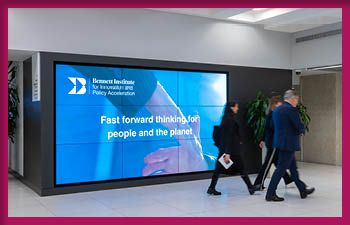 A large digital screen in a modern building displays an advertisement for the Bennett Institute for Sustainable Public Policy Acceleration. The screen shows the institute's logo and the text 