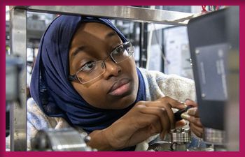 A young woman wearing a navy blue hijab and glasses works intently with delicate mechanical equipment, adjusting components with her hands in a laboratory or technical setting.