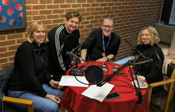 A photograph of four people sitting at a round table, which has microphones and other podcast equipment on it