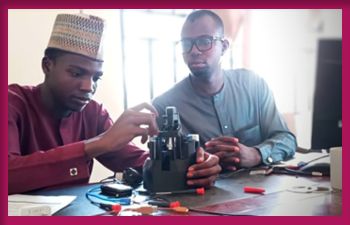 Two men, one wearing a maroon traditional outfit and hat, and the other in a blue-grey traditional outfit with glasses, work together on assembling a biomedical device at a desk.