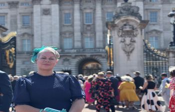 A smiling Lisa Holmes stands in front of the gates of Buckingham Palace. One can see other garden party guests arriving in the background.