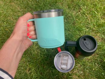 Three different reusable mugs - one is being held - with grass as the backdrop