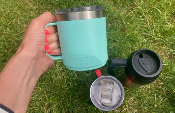 Three different reusable mugs - one is being held - with grass as the backdrop