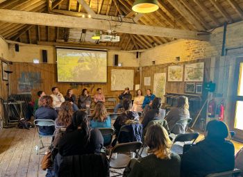 A group of ten students sit in a large wooden room listening to a panel of seven Knepp staff members. The live camera of a Stork nest is displayed on a projector behind the panel.