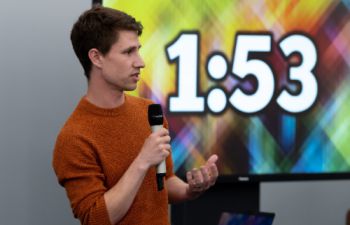3MT presenter holding a microphone with a timer in the background