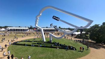 Large sculpture of a face with cars inside it at the Festival of Speed, with crowds and tents in the background.
