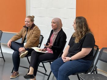 Three diverse women sit on chairs, in discussion with an unseen audience.