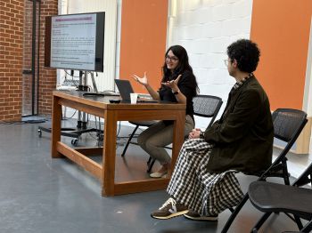 Two women sit on chairs, one smiling and the other talking, in discussion with an unseen audience.