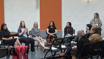 Five diverse women sit on chairs in front of an audience, one of them speaking animatedly. Another woman, the chair of the panel, stands nearby and listens.