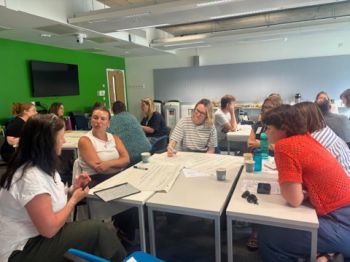 A group of people seated around tables in a classroom setting, engaged in collaborative discussions with papers and writing materials. A green wall with a mounted TV screen is visible in the background.