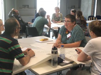 A group of people are sitting around tables in a classroom or meeting room setting. They are engaged in discussions and writing on large sheets of paper. Some individuals have name tags, and there are various items such  notebooks, and pens