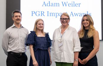 Four post-graduate researchers standing together and smiling with a screen behind them displaying the text 