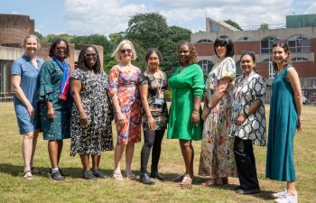 A group of nine women standing in a row on a sunny day at the University of Sussex campus, smiling at the camera.