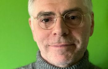 Close up of Paolo Belloni in front of a green background. Paolo has glasses and wears a turtleneck jumper.