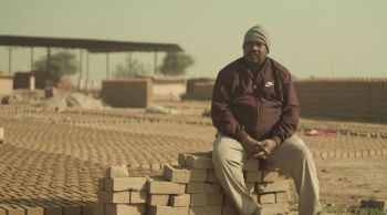 In this still from Pratik's documentary film, an Asian man sits atop a pile of bricks in a sandy, rather desolate landscape.